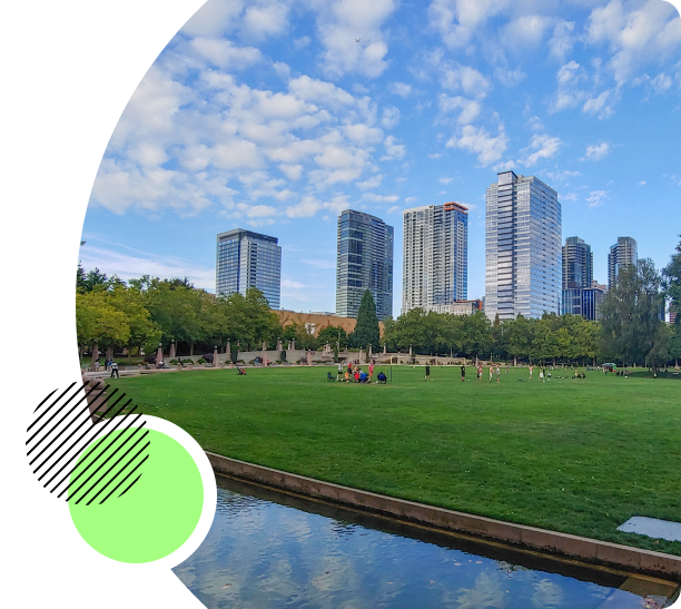 Scenic view of downtown Bellevue, featuring modern skyscrapers against a blue sky with scattered clouds, a large green park in the foreground with people enjoying the open space, and a calm water feature reflecting the sky.